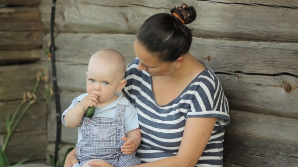 Beautiful Baby Boy Sits At Mum On Hands And Eat Cucumber. Near An Old Wooden House