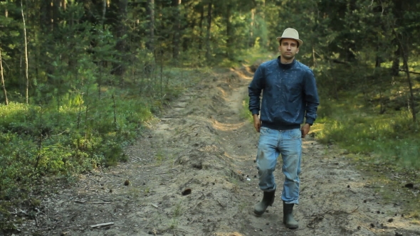 Young Hiker Man With Hat Walking In Forest. He Adjusts His Old Jeans And Goes On