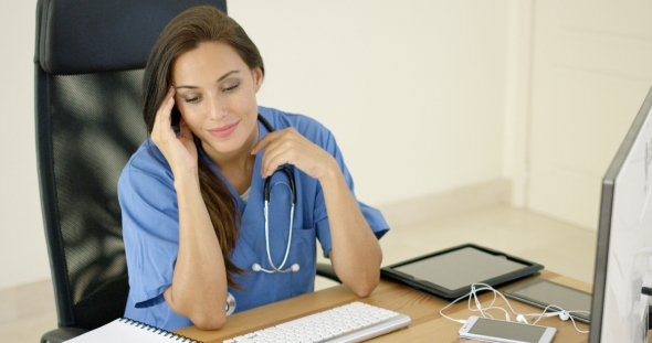 Healthcare Worker Wearing Scrubs Sits At Computer alt