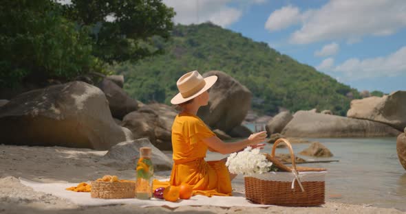 Woman in Straw Hat and Yellow Dress Holding Glass of Water Sitting on Lonely Beach on Summer Day alt