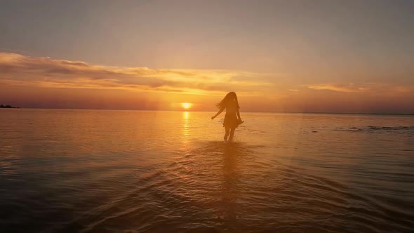 Brunette woman in a dress running along water at sunset or sunrise alt