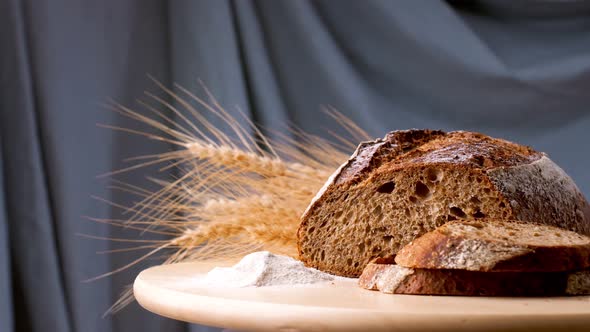 Dark Bread on a Wooden Board with Ears of Wheat and Flour Rotates on a Gray Background Closeup alt