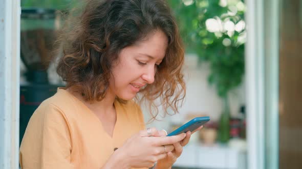 Smiling Curly Woman in Yellow Dress Using Her Phone Happily While Sits in Coffee Shop alt
