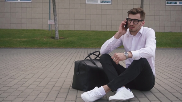 Handsome Young Businessman In Glasses Talking On The Phone Outdoors While Sitting On The Ground At