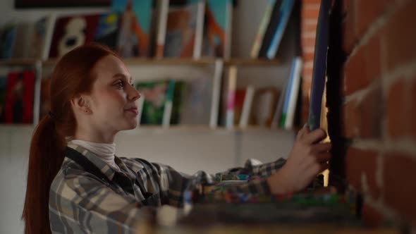 Side View of Happy Smiling Female Artist Putting Finished Modern Painting on Canvas on Shelf in alt