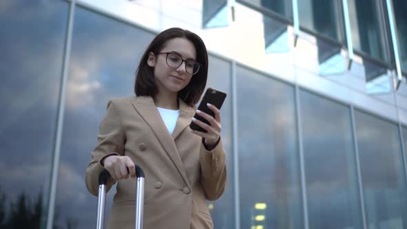 A Young Woman Stands with a Phone on the Background of a Business Center. Girl in a Coat with a alt