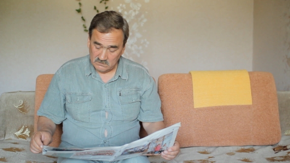 Senior Man At Home Reading Newspaper And Sitting On Sofa
