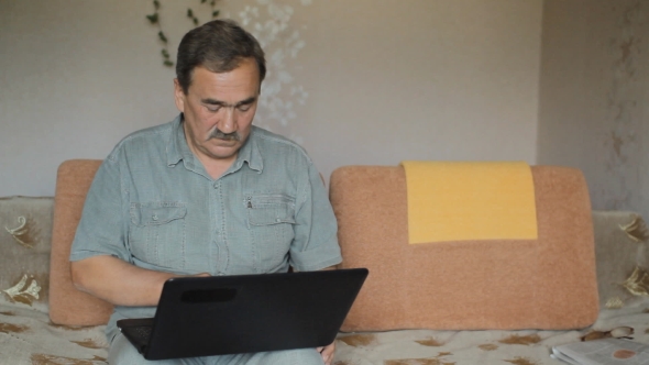 Senior Man Sitting On Sofa With Laptop At Home. He Carefully Works At The Computer.