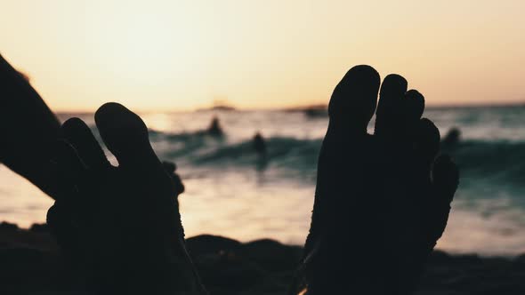 POV Silhouette of Feet of Young Man Lying on Sandy Beach By Ocean During Sunset alt