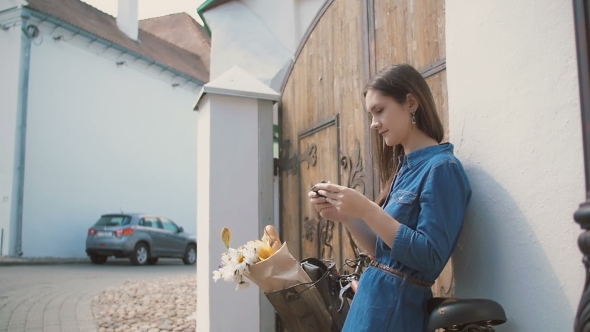 Brunette Girl Using Her Smartphone Standing Near Building With a Bike With Flowers In a Basket,  alt