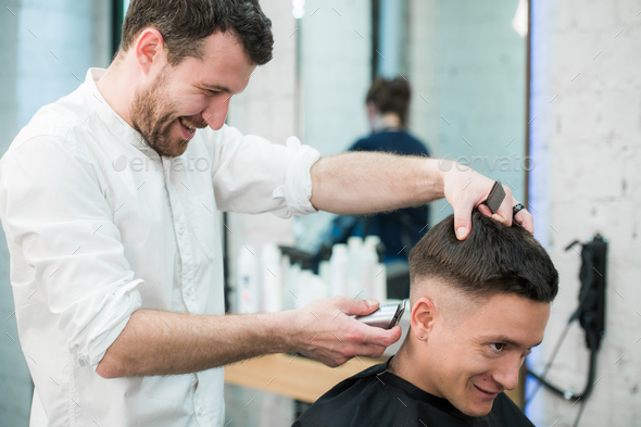 Professional styling. Close up side view of young satisfied man getting haircut by hairdresser with