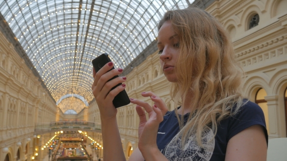 Pretty Young Blonde Is Using Smartphone In Background Of Illuminated Roof And Walls Of Mall