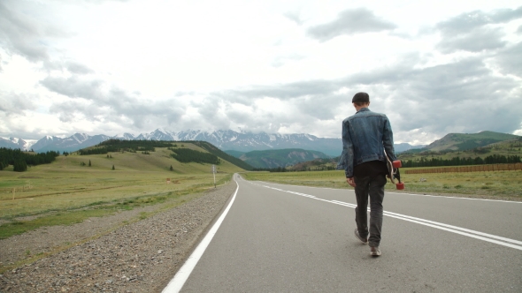 Man Carries a Longboard. The Boy Goes On The Asphalted Road Holding a Longboard alt