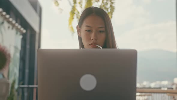 A Young Asian Girl Works at Her Laptop in a Rooftop Cafe in the City alt