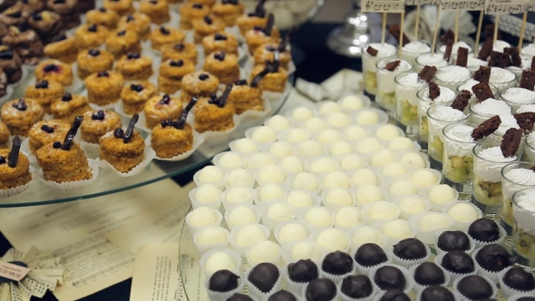 Banquet Table And Laid Out a Variety Of Small Cakes, Candies, White And Dark Chocolate And a Glass alt