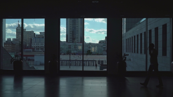 Man Goes Dark Corridor Of Office Space With Large Windows. alt