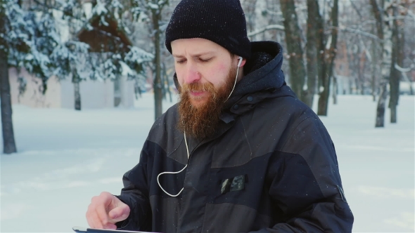 Steadicam Shot: Attractive Bearded Man Walking In a Winter Park, Enjoys a Tablet alt