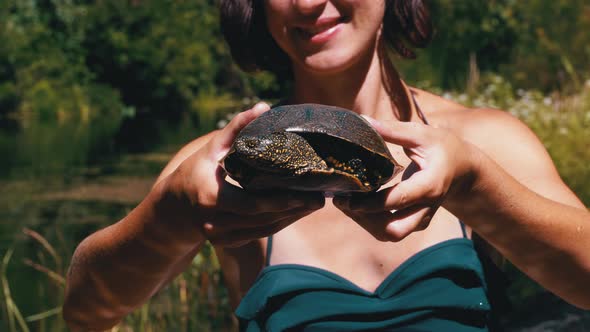 Woman Holds Funny Turtle in Arm and Smiles on River with Green Vegetation alt
