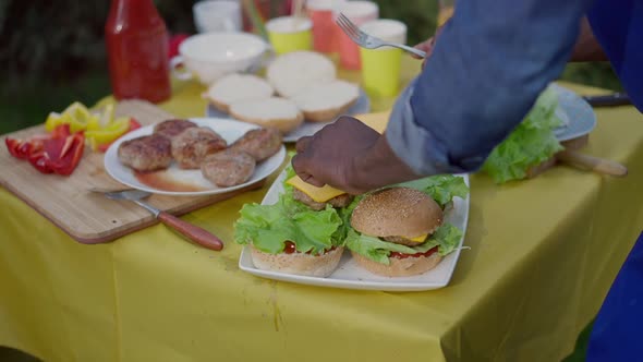 Unrecognizable African American Man Making Delicious Meat Burgers on Table for Picnic alt