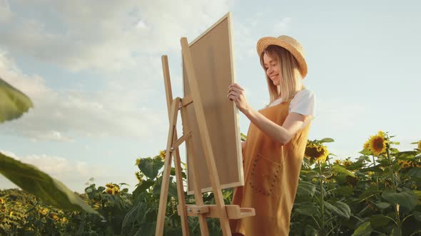 A Young Woman is Putting the Painting on an Easel alt
