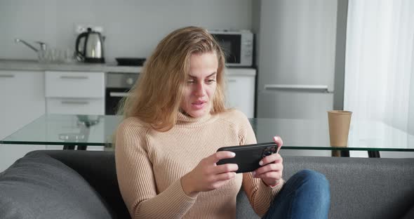 Blonde Millennial Woman in a Beige Woolen Sweater Play Game with Smart Phone on Modern Kitchen alt