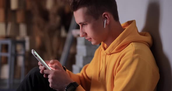 Man in Yellow Hoodie Using Smartphone and Earbuds at Home alt