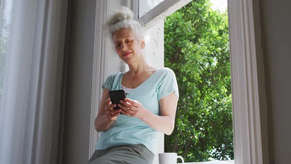 Senior caucasian woman using smartphone near the window at home alt