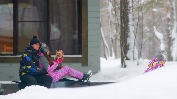 Man Hugs Wife with Dog While Daughter Plays with Snow alt