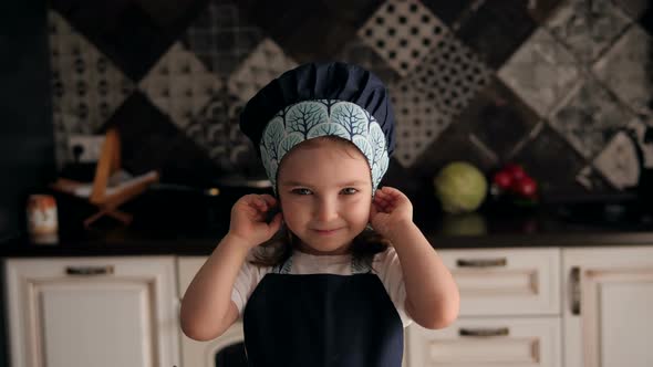 Portrait of a Little Charming Girl of 4 Years in a Cap and Apron in the Kitchen alt