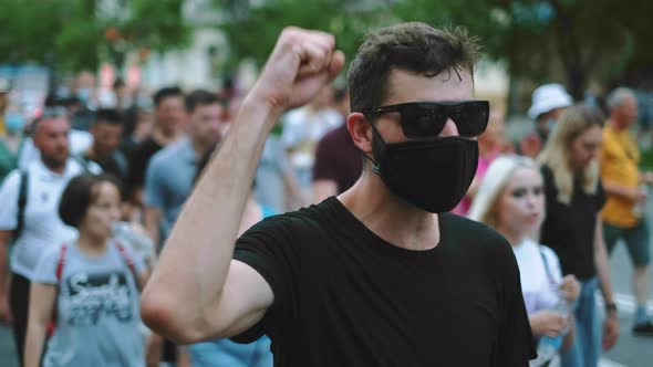 Protester Guy in Black Eyeglasses and Face Mask on Riot Crowd Waves Arm Fist