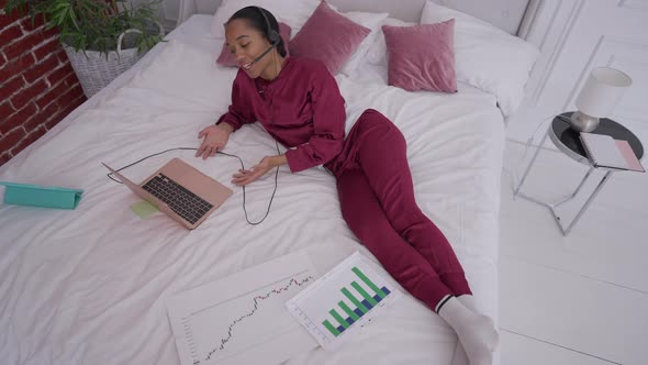 Top View of Joyful Slim African American Young Woman Lying on Bed Conferencing Online on Laptop alt