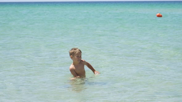 Blond Boy is Learning to Swim in the Clear Azure Sea on a Sunny Day