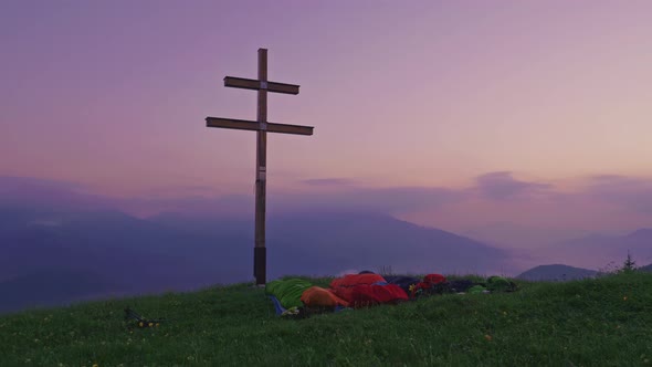 Group of people wild camping on the mountain peak with cross and green meadow. People in sleeping ba alt