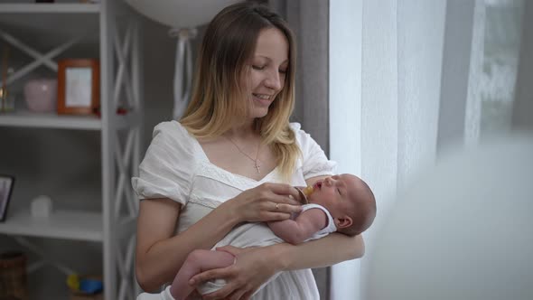 Smiling Woman Standing at Window in Living Room Putting Pacifier in Mouth of Newborn Baby Shaking alt