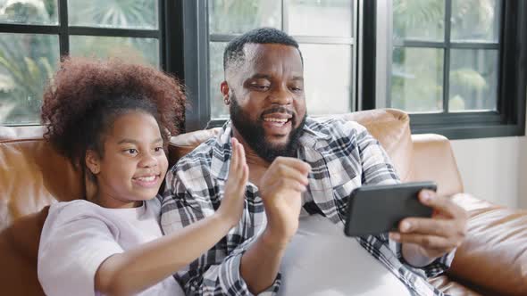 Happy African family dad and daughter having fun and using mobile phone video call on sofa at house alt