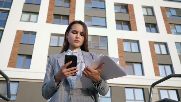Young Woman in a Business Suit Looks at the Phone and at the Documents on the Background of the alt