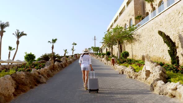 Back View of Young Woman in Summer Clothes and Sun Hat, Walks with Travel Luggage Along an Empty alt