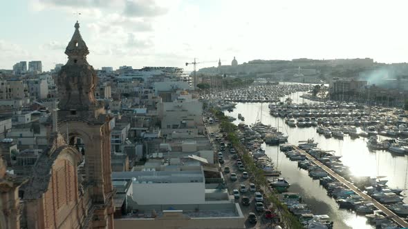 Small Village in Malta Revealing Behind Two Church Bell Towers with Boats in Port on Sunny Day alt