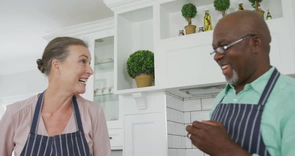 Smiling senior diverse couple wearing blue aprons and cooking in kitchen alt