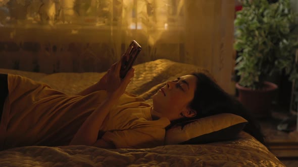 Pretty young caucasian woman is lying on back on the bed, and using a smartphone. alt