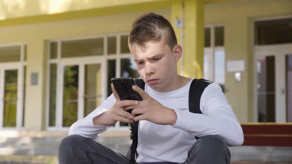 A Caucasian Teenage Boy Works on a Smartphone As He Sits in Front of School  Closeup