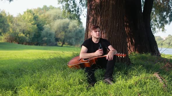 A Young Guy Sits Under a Tree in His Hands Holds a Guitar and Drinks a Bottle of Beer