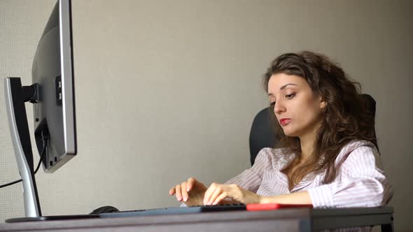 Tired Young Woman with Curly Hair and White Shirt is Working at the Office Using Her Computer alt