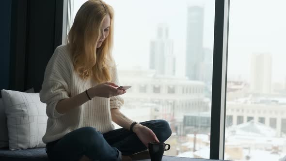 smiling woman uses smartphone while sitting on the windowsill with city view