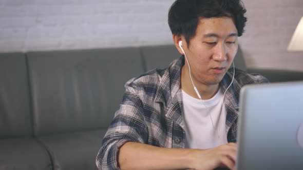 Young Asian Male Entrepreneur in Casual and Earphone Work on Computer Laptop at Late Night From Home alt