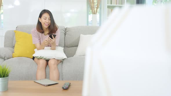 Young Asian woman using smartphone checking social media feeling while lying on the sofa when relax.