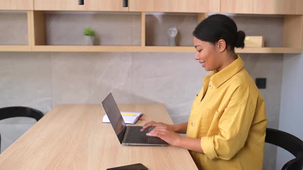 Smiling Young Multiracial Female Entrepreneur Sitting at the Table with Laptop in the Kitchen