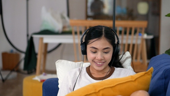Portrait a beautiful young Asian woman while on a sofa in headphones listening music. alt