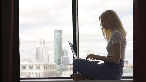 Business Woman Sits on the Windowsill and Types Text on the Laptop Keyboard