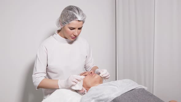 Doctor Beautician Removes Makeup Patient Lying on a Special Table alt